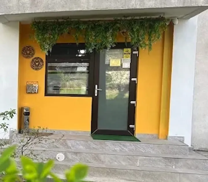 Intrarea în salon: ușă cu tâmplărie verde într-un cadru galben, la parterul unui bloc, cu verdeață deasupra